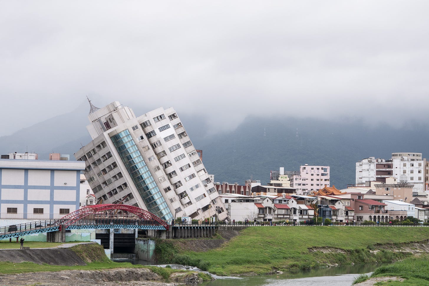 Prédio tombado após terremoto. Dia nublado e prédio tombado à esquerda ao lado de casas e às margens de um rio