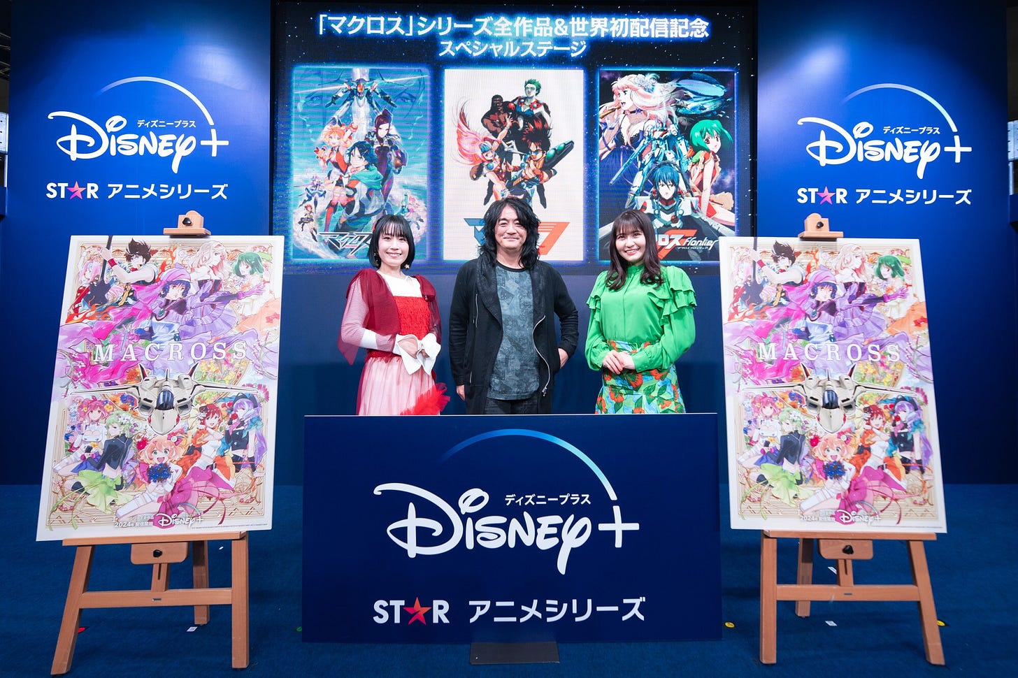 A man and two women are flanked by two large posters of the Macross anime franchise standing on easels. In front and behind them are banners displaying the logo of the Disney+ streaming service. On the projection screen behind them, posters from three different Macross titles are also displayed.