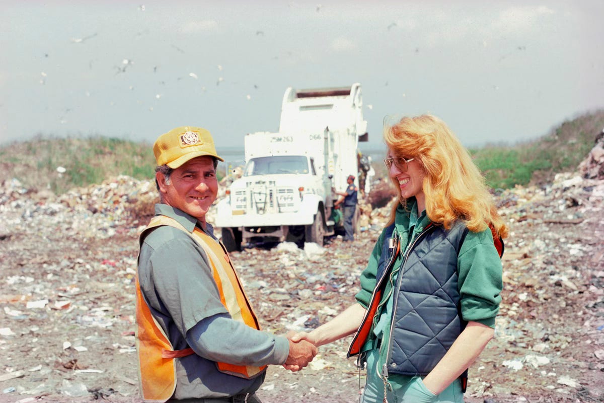 A photograph of two people shaking hands. On the left is a white male sanitation worker in a safety vest, looking at the camera and smiling. On the right is a white woman, Mierle Laderman Ukeles, smiling warmly and happily at the sanitation worker. She has cascading red-gold hair, and is wearing a blue quilted vest over a green sweatshirt. In the background is a garbage dump site with mounds of garbage receding into the distance. There is a big white sanitation truck emptying its load. Above a flock of seagulls catches the sunlight. A photograph of two people shaking hands. On the left is a white male sanitation worker in a safety vest, looking at the camera and smiling. On the right is a white woman, Mierle Laderman Ukeles, smiling warmly and happily at the sanitation worker. She has cascading red-gold hair, and is wearing a blue quilted vest over a green sweatshirt. In the background is a garbage dump site with mounds of garbage receding into the distance. There is a big white sanitation truck emptying its load. Above a flock of seagulls catches the sunlight.