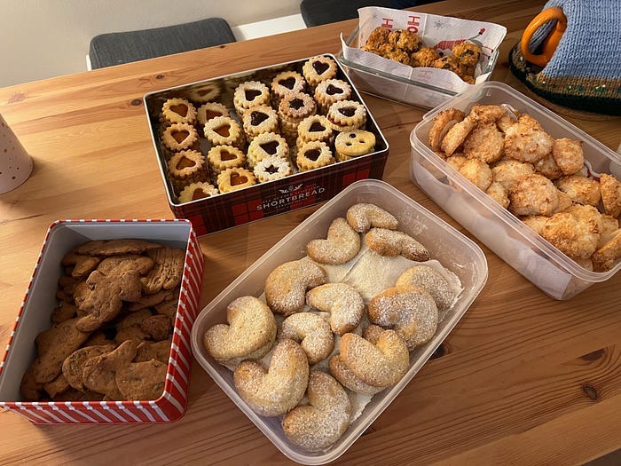 5 tins of home made Christmas Cookies next to a teapot that is under a knitted warming hood