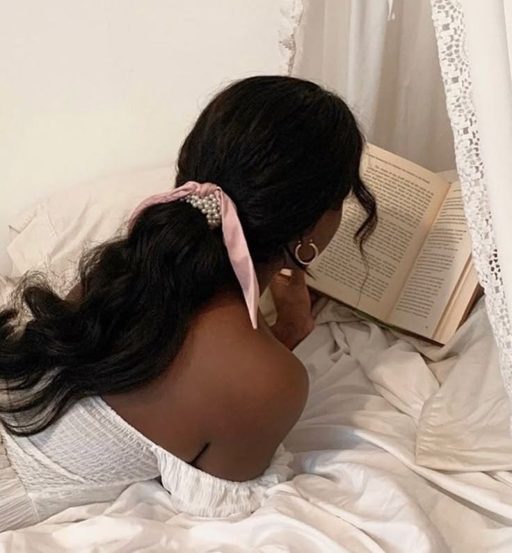 This contains: dark skinned girl with hair in a ribbon ponytail reading a book on a white bed with a white canopy