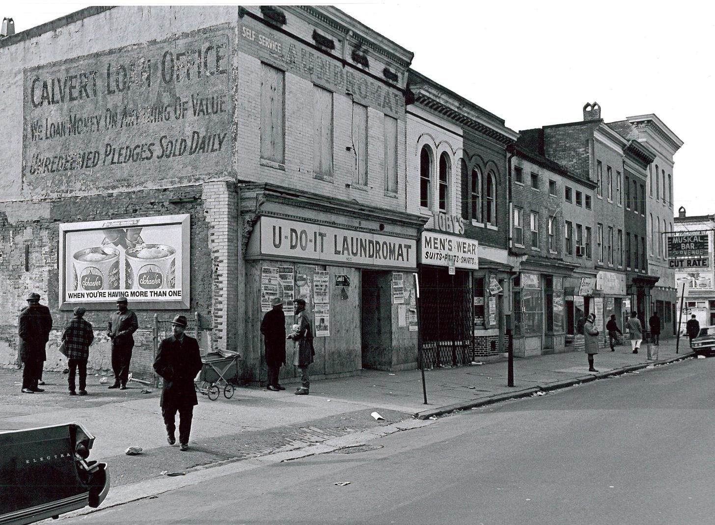 Black-and-white photograph of a stretch of Baltimore City storefronts showing significant impacts of the 1968 unrest. Many are boarded up and all appear closed. People walk by on the street.