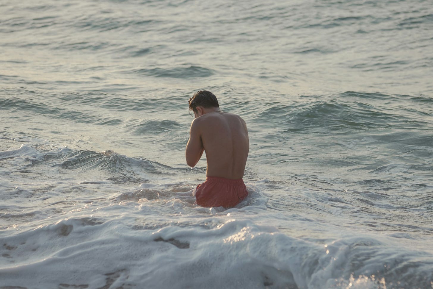 Un ragazzo con costume bermuda arancione di schiena in piedi in mezzo al mare, mentre le onde in primo piano fanno schiuma. Il sole arriva con dei riflessi morbidi, come se fosse l'ora del tramonto. Il ragazzo si porta le mani al viso.