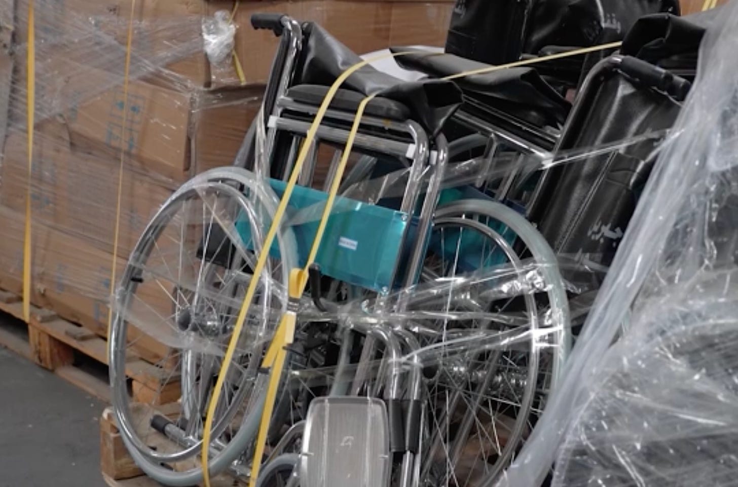 A still from video showing several manual hospital-style wheelchairs tied and taped together in a storage facility.