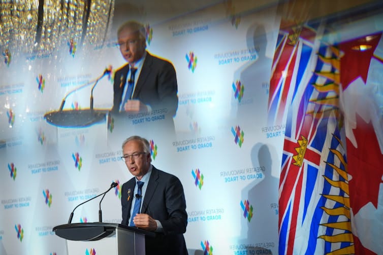 A grey-haired man with glasses is reflected in mirrors as he speaks from behind a lectern. A red blue and white flag is to his right.