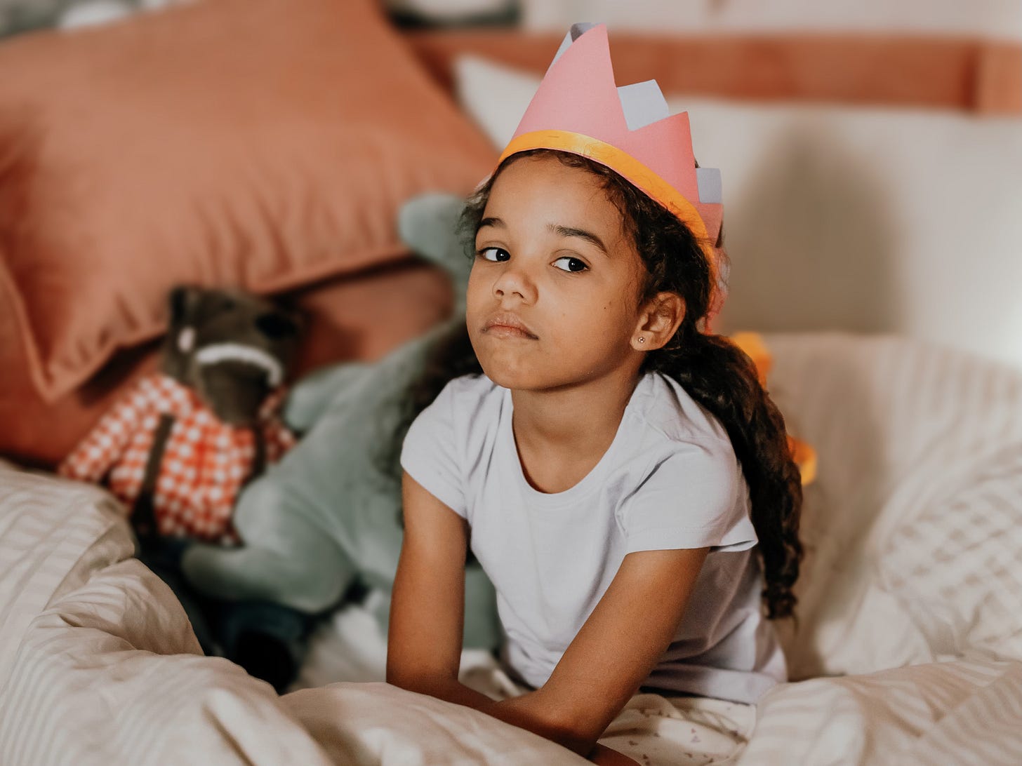 Photo of a sad biracial girl sitting in her bed. She has a paper crown on her head.