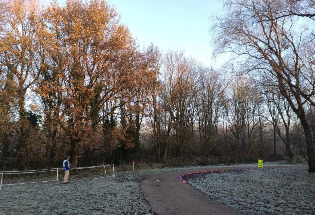 The sun lights the leaves left on the trees, overlooking the run director at the finish line, with pink and orange cones keeping runners out of the mud just off the path.