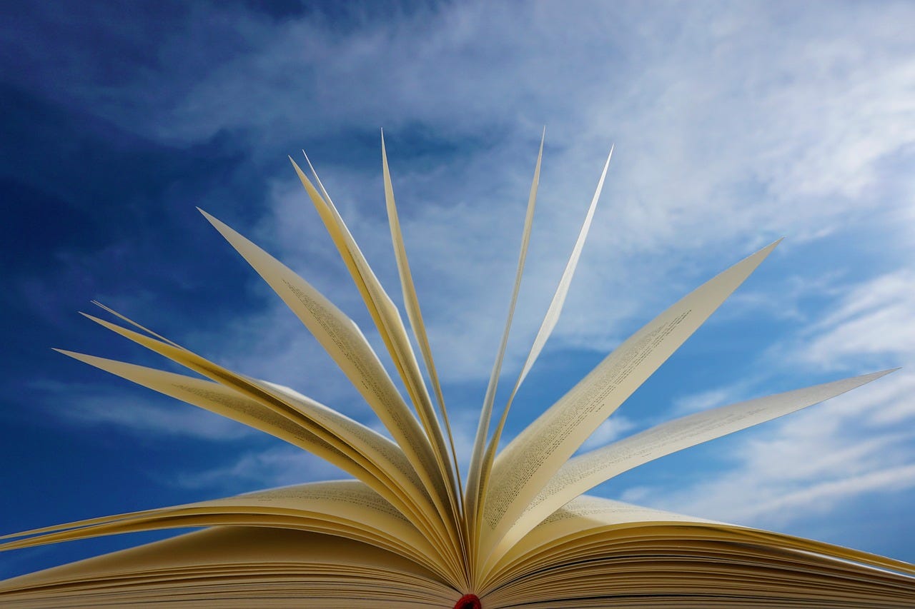 A book with pages fanned out in front of a blue sky with clouds wafting by