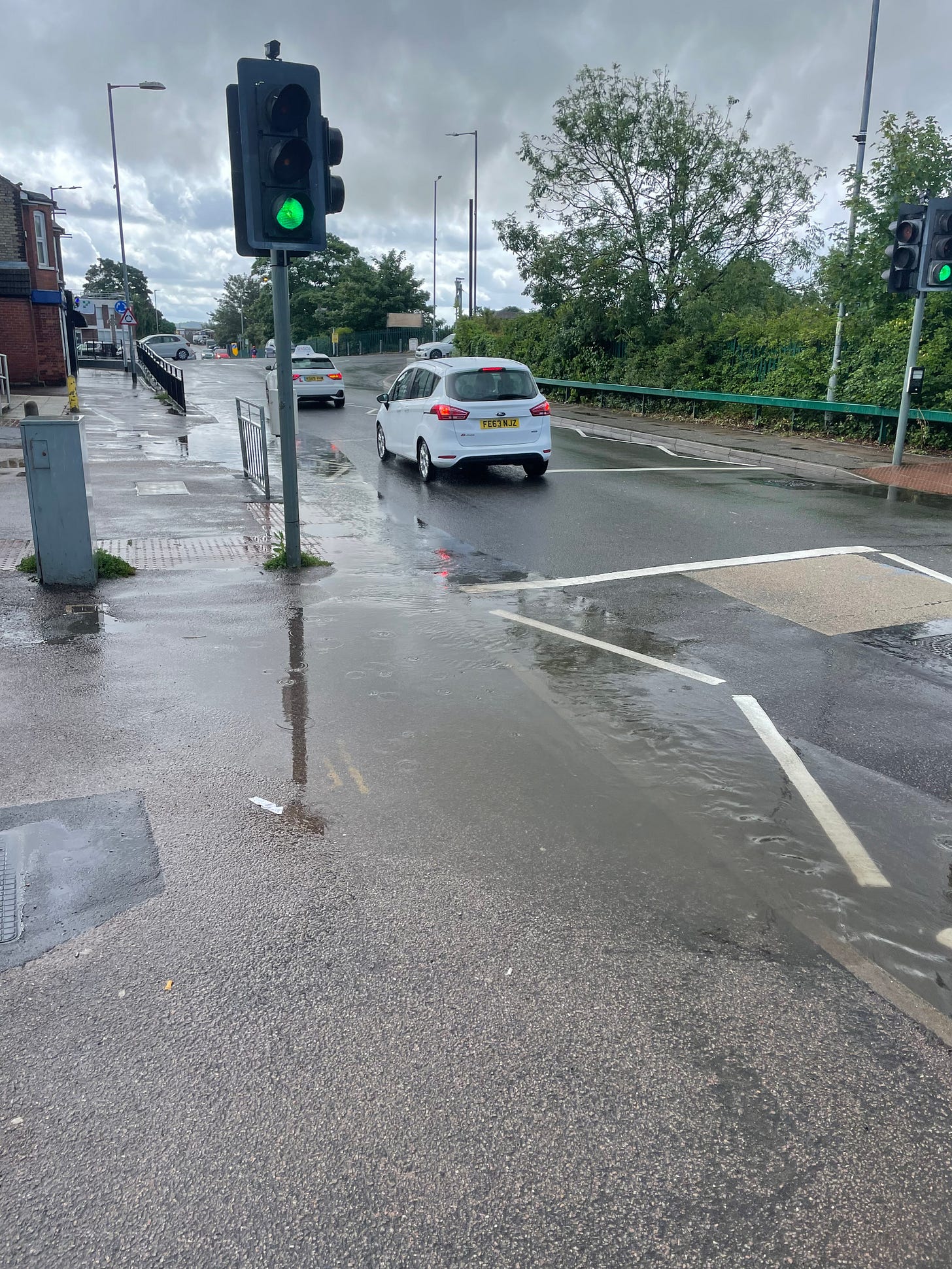 a pool of water sits across a pavement and pedestrian crossing. the lights are green, and cars are passing