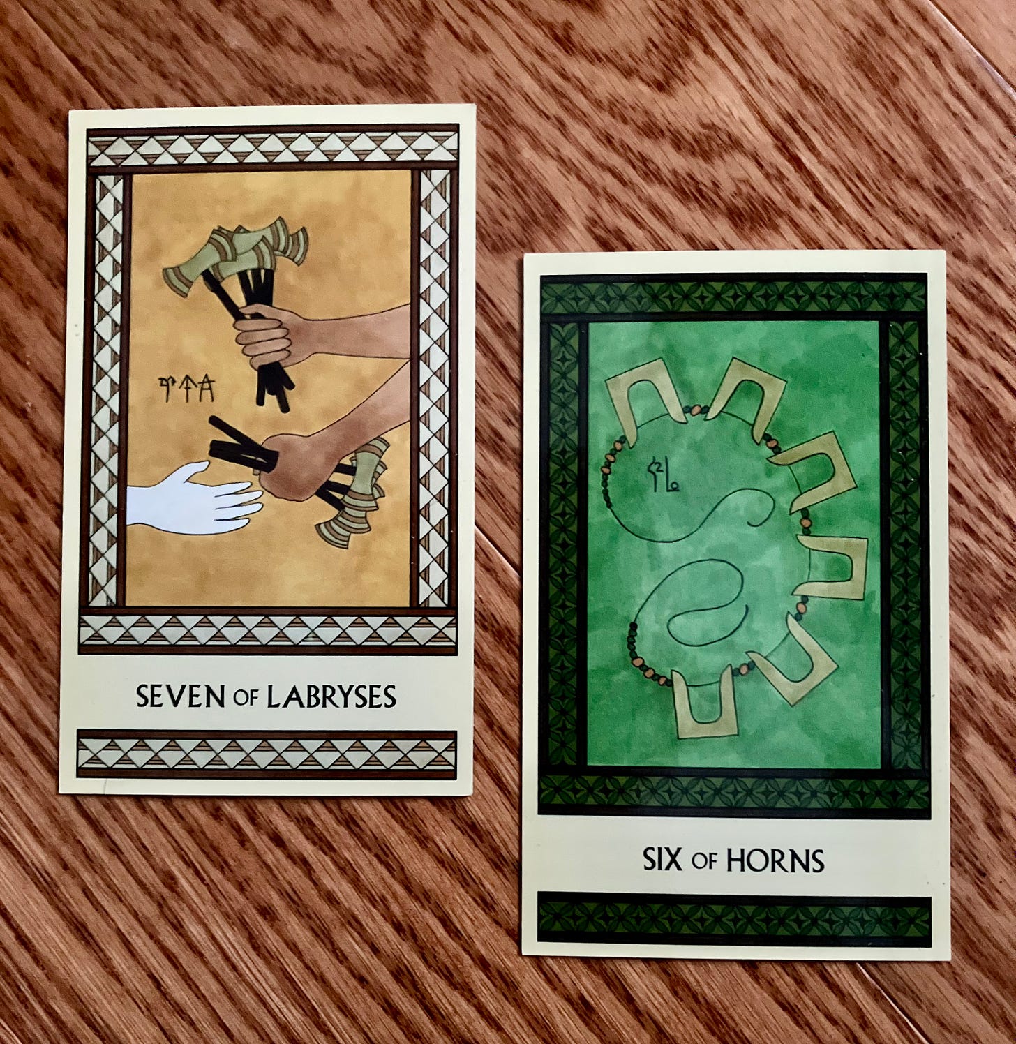 Two Minoan Tarot cards side by side on a wood background. The Seven of Labryses is in shades of gold and cream and shows one set of hands holding a bunch of labryses and handing some of them to another. The Six of Horns is in shades of green and shows a necklace made of six pairs of golden sacred horns.