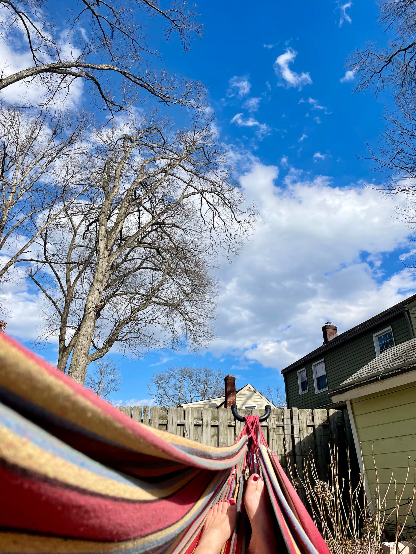 A red and yellow striped hammock against blue sky with white clouds and bare winter trees.