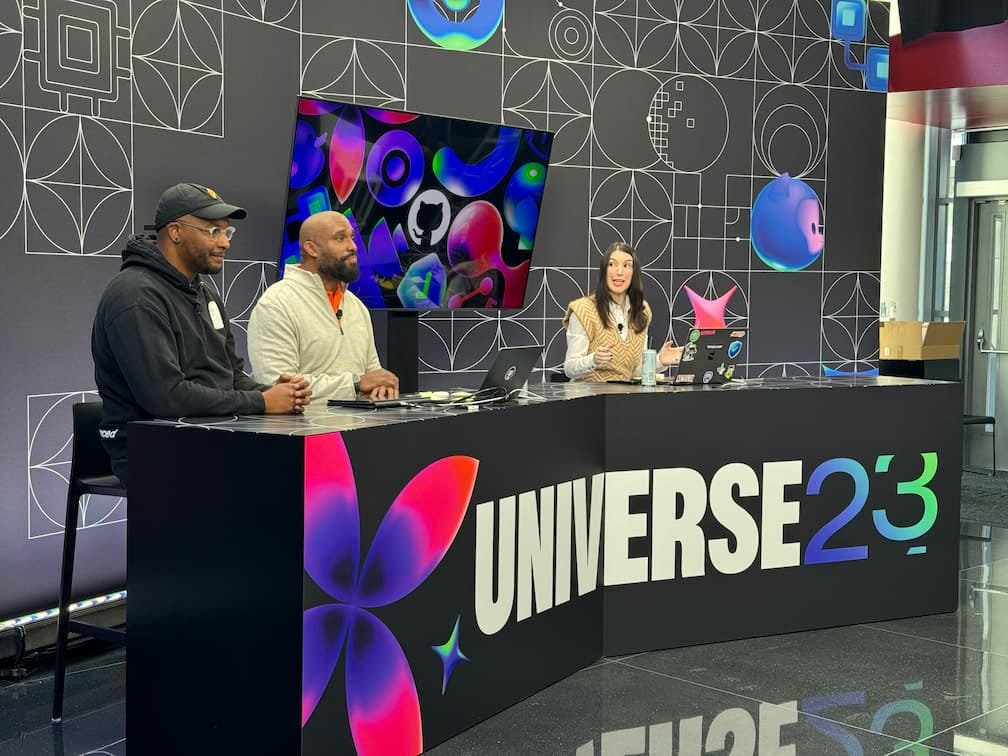 A photo of a TV news style desk carrying the logo Universe 23, with three people sat behind it.