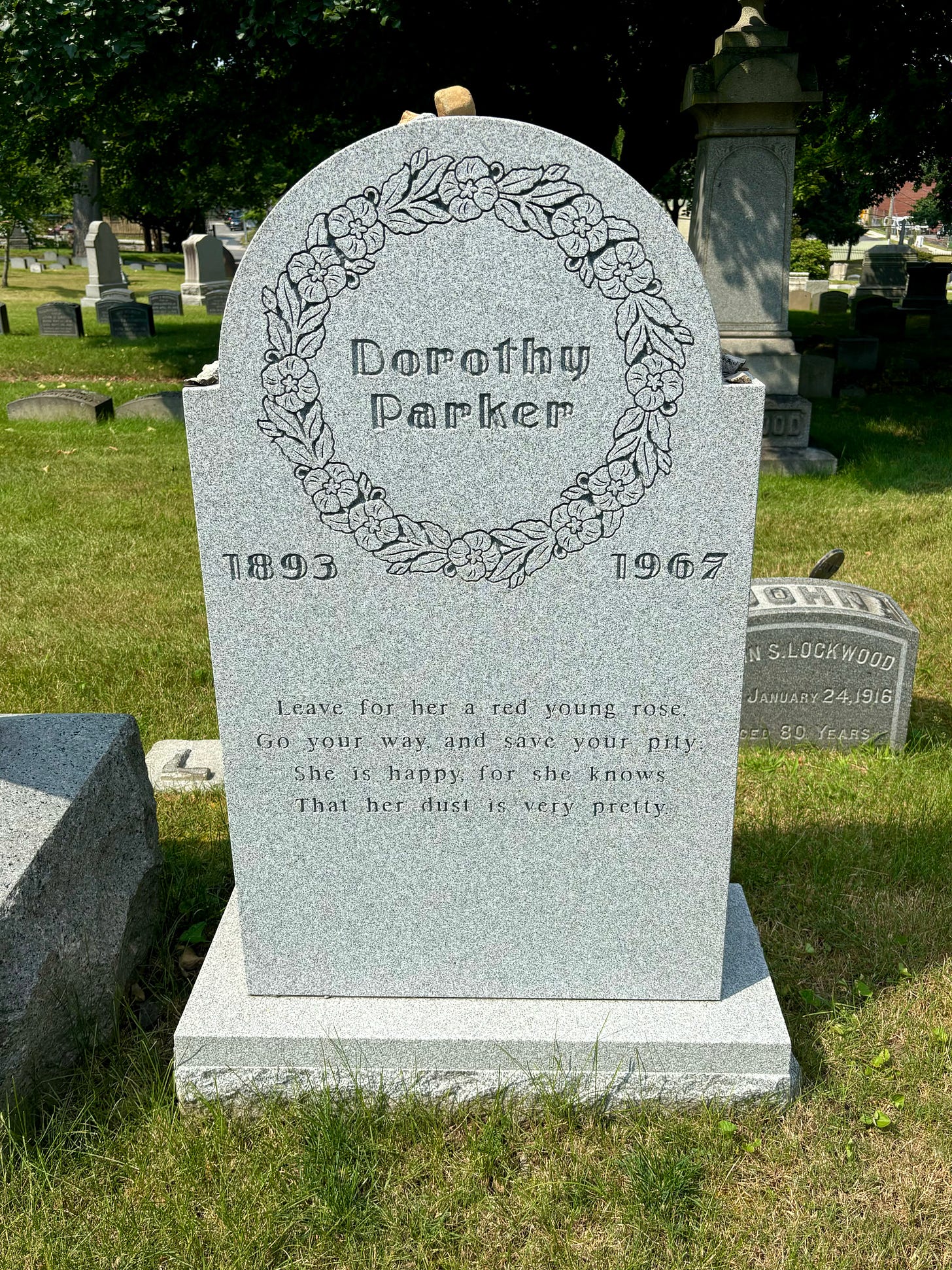 Dorothy Parker's headstone. It is sizable but tasteful. Her name is surrounded by a carving of a wreath, echoing the rounded top. It has her birth and death years below and on either side of the wreath around her name, and the epitaph is below. On top are stones placed by mourners. The sun is shining and the grass around her is worn but green.