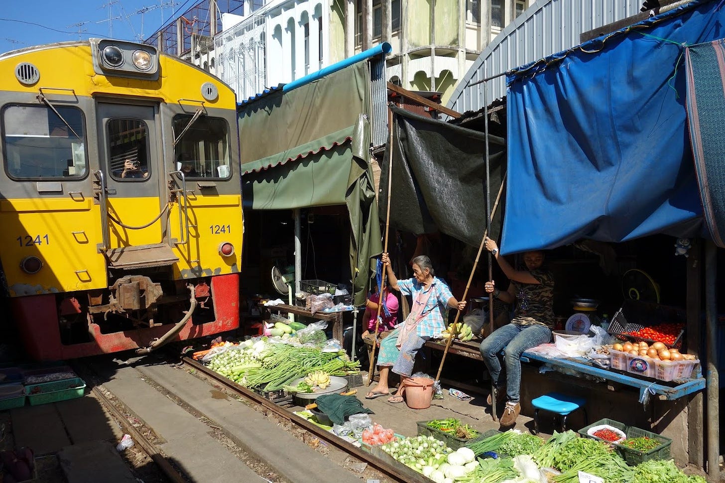 A railway with a train passing extremely close to market shops. The train passes OVER produce arrayed for the market and the shops have folded in their canopies which just barely clear the train. Shopkeepers sit on benches watching the train pass within feet of them.