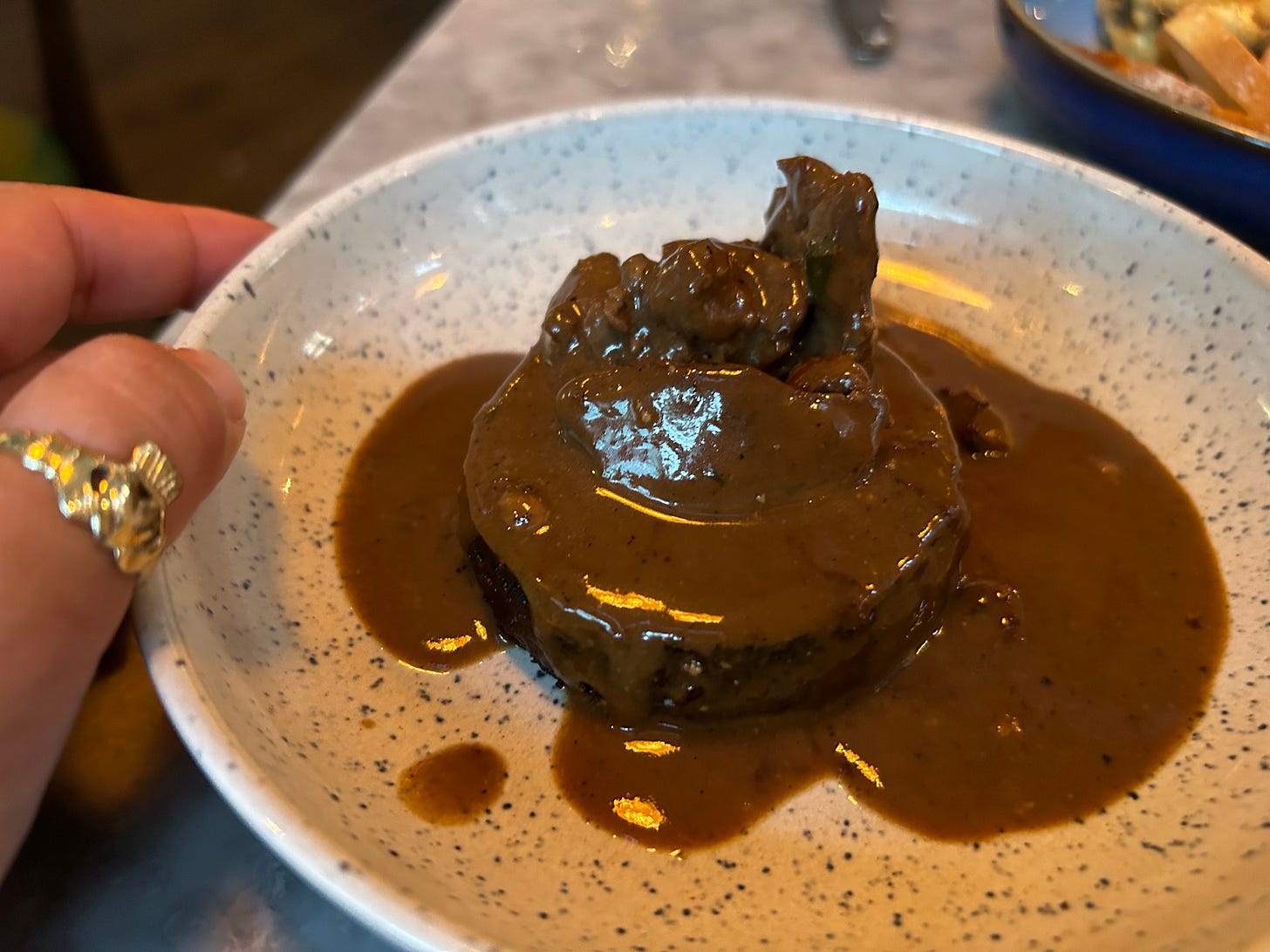 A hand wearing a claddagh ring, holding a plate of black pudding and chicken livers, with gravy poured on top.