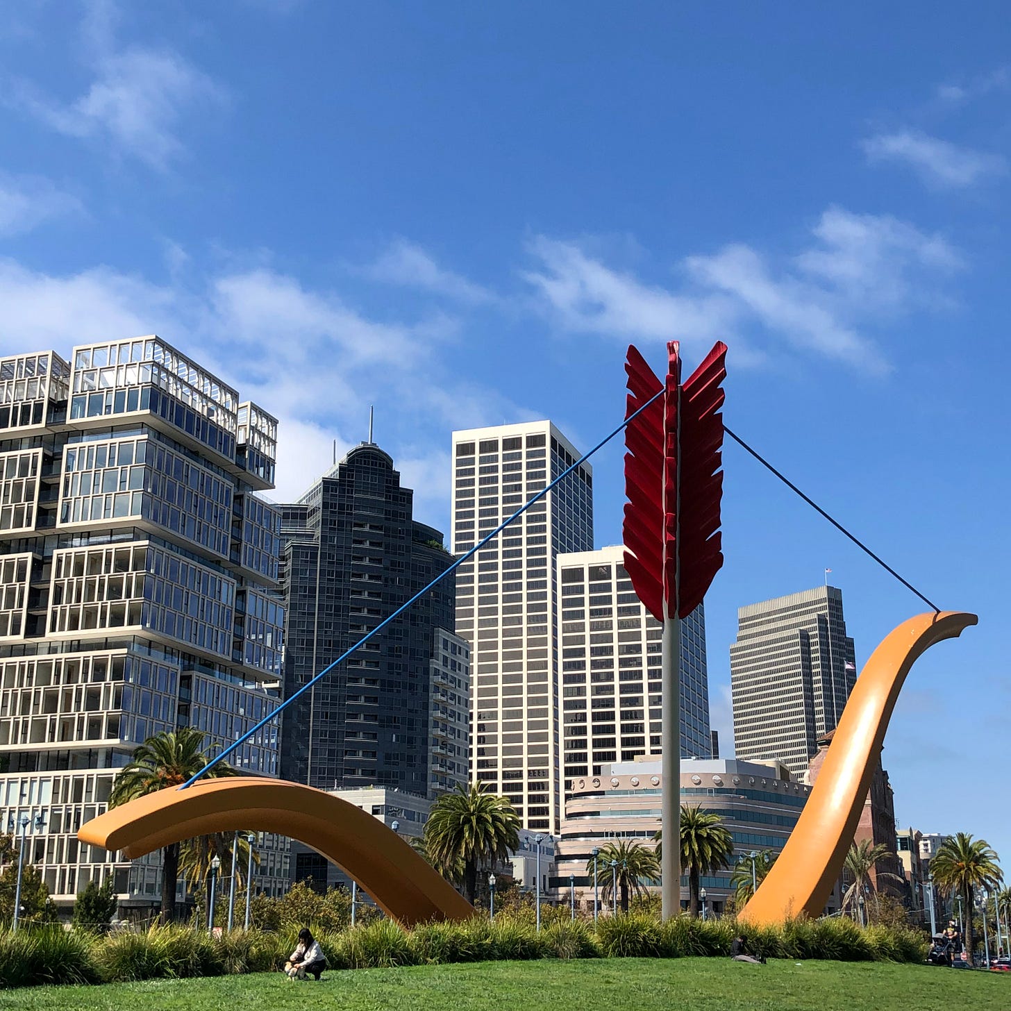 Giant bow and arrow with the arrowhead implanted into a grassy knoll, city skyline in the background, bright blue sky. Petite woman kneeling in front of the bow and arrow sculpture