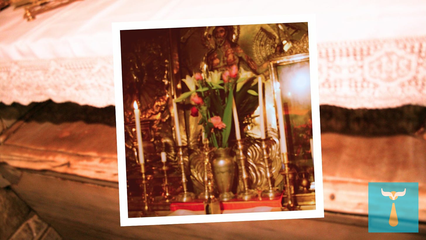 A Polaroid style photograph of a brilliantly decorated altar with many tall candlesticks and pink and red roses with white lilies also shows a painted Risen Christ on metal artwork behind the altar. Behind the Polaroid is a slightly blurry photo of a stone-covered ledge and a shelf-style altar above it covered with a white lace altar cloth.