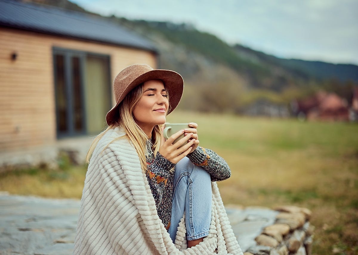 Photo of a smiling young woman, eyes closed, seated ouside a cabin, holding a cup of coffee, wearing a hat, wrapped in a blanket