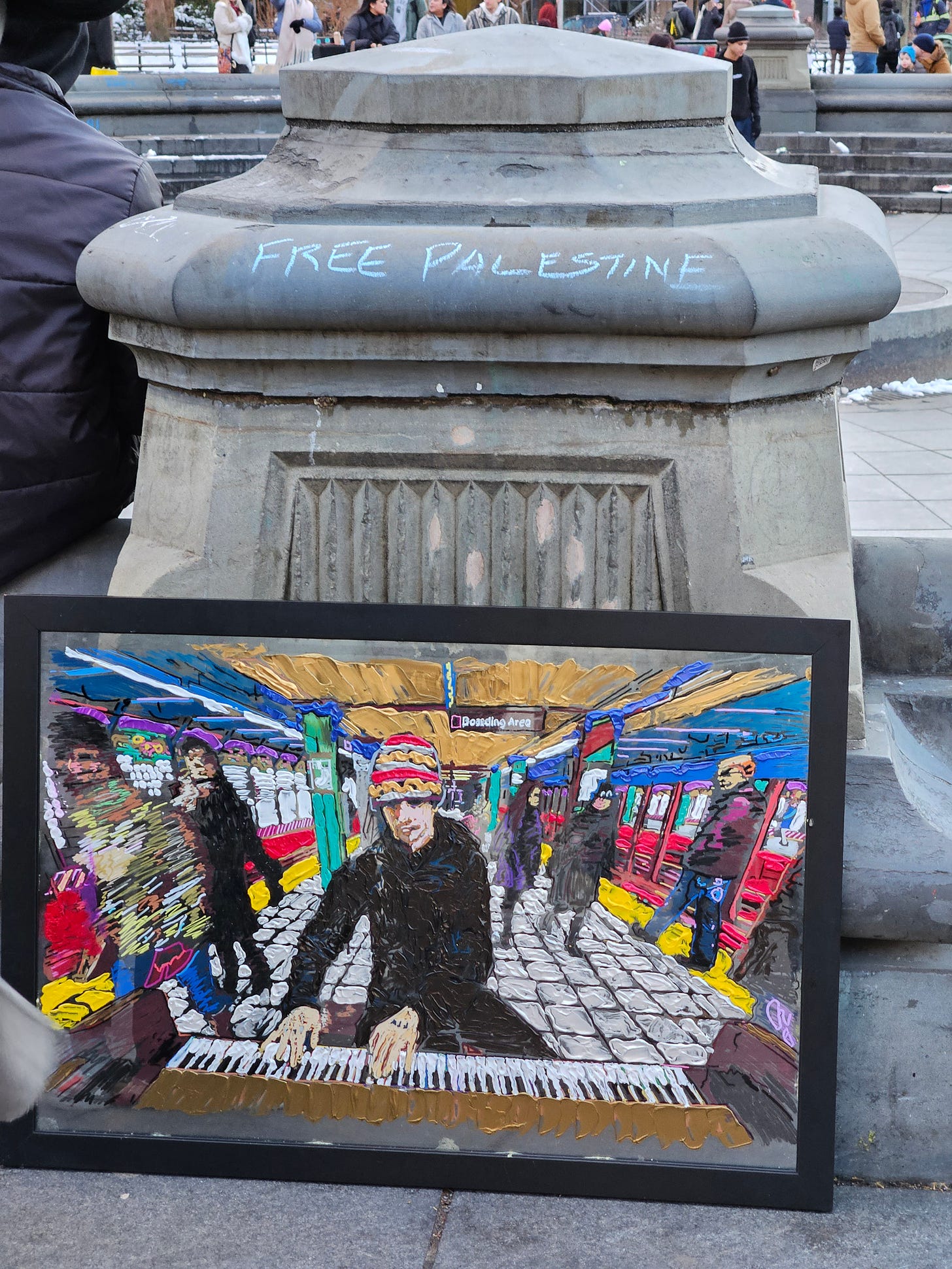 "Free Palestine" is written in blue chalk across the top of a cement pillar. A painting is leaned against the pillar that shows a colorful depiction of a pianist playing on a subway platform. 