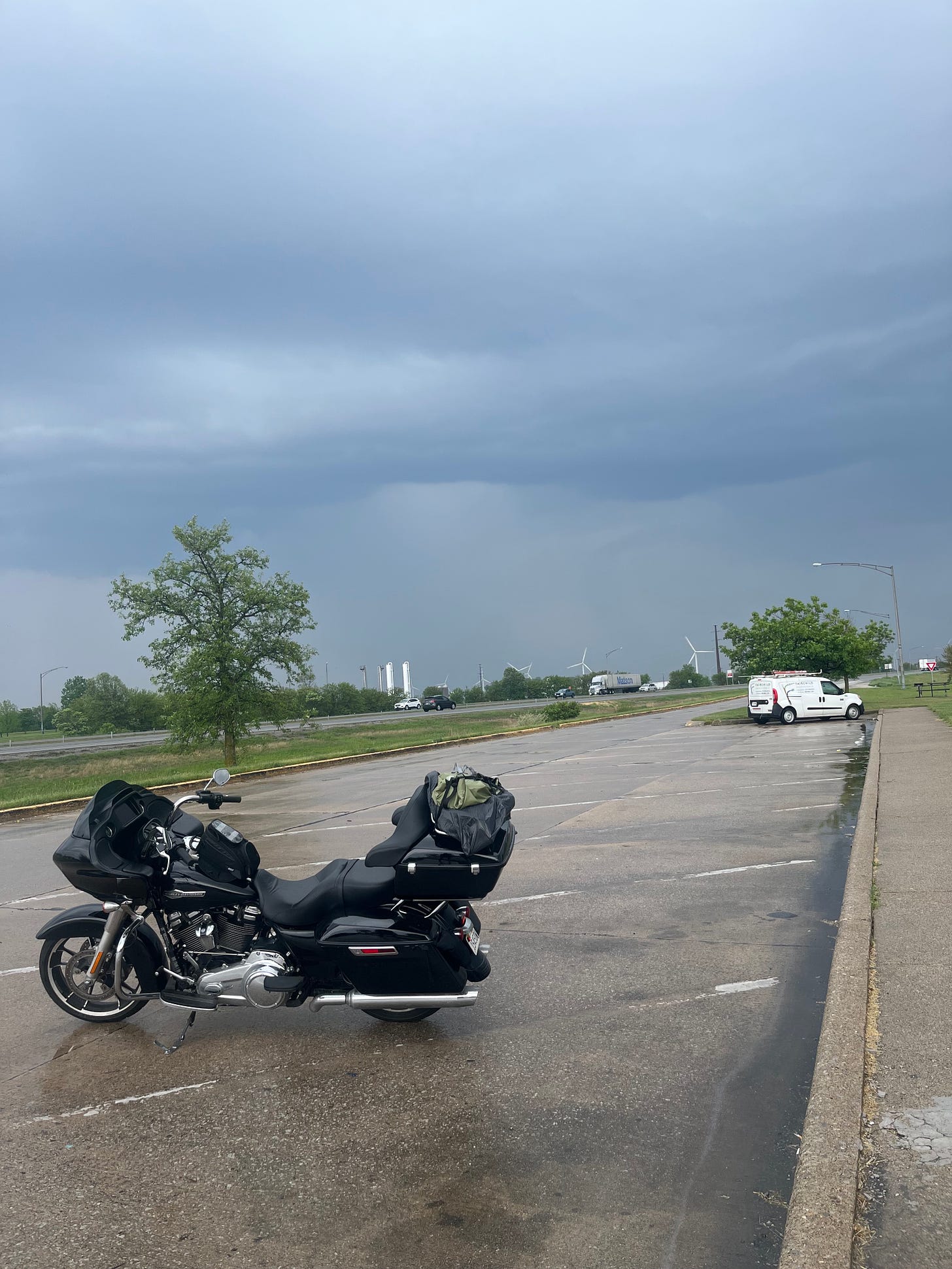 The motorcycle parked in a parking lot off the freeway with a cloudy and dark sky overhead. 