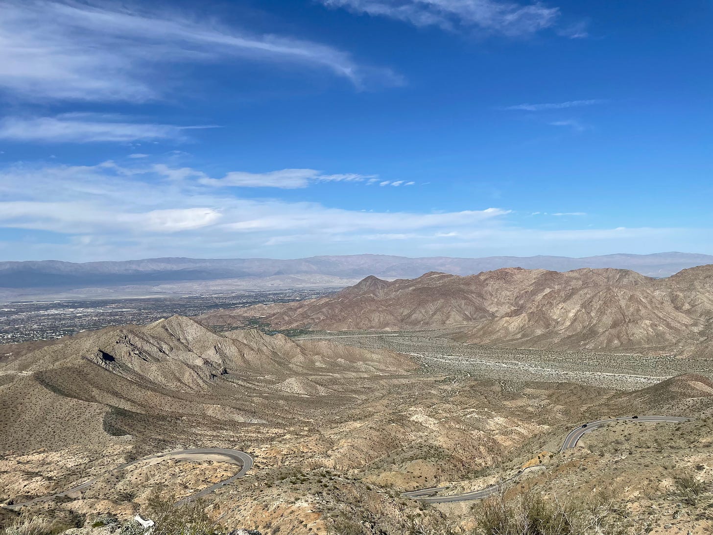 View, from above, of the Coachella Valley, a desert-like expanse scattered with mountains. The sky is blue with sparse clouds.