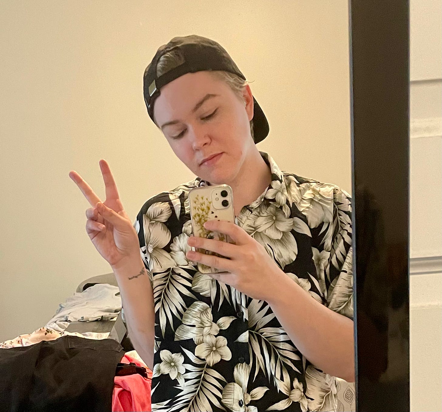 mick stands in front of a mirror taking a photo. they are wearing a black baseball cap backwards and a black and white Hawaiian shirt. they are holding up two fingers to make a peace sign.