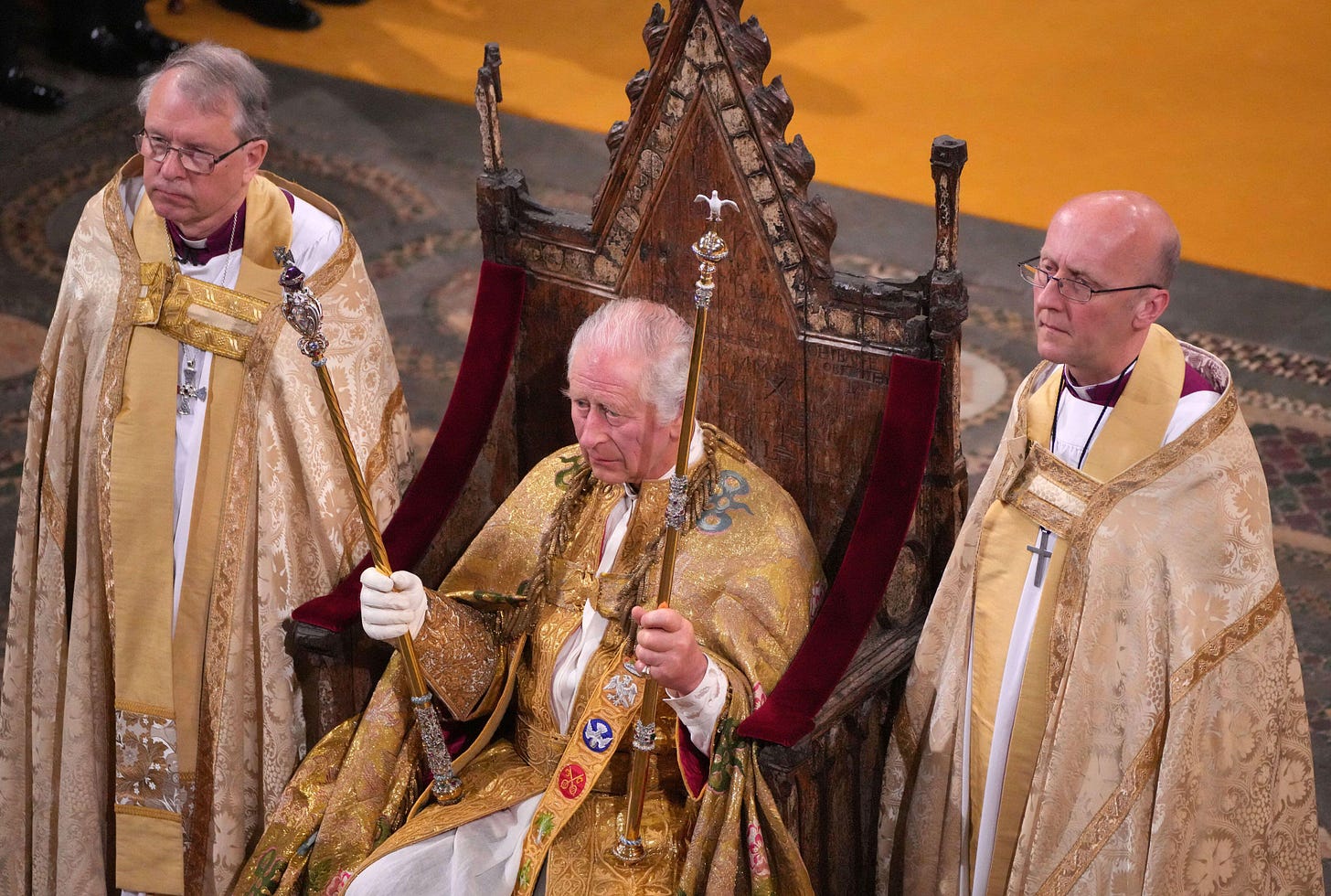Coronation of Kings Charles III on 6 May 2023. Charles is sitting in the center holding two scepters from the Crown Jewels and is flanked on either side by priests. Everyone is dressed in white and gold.