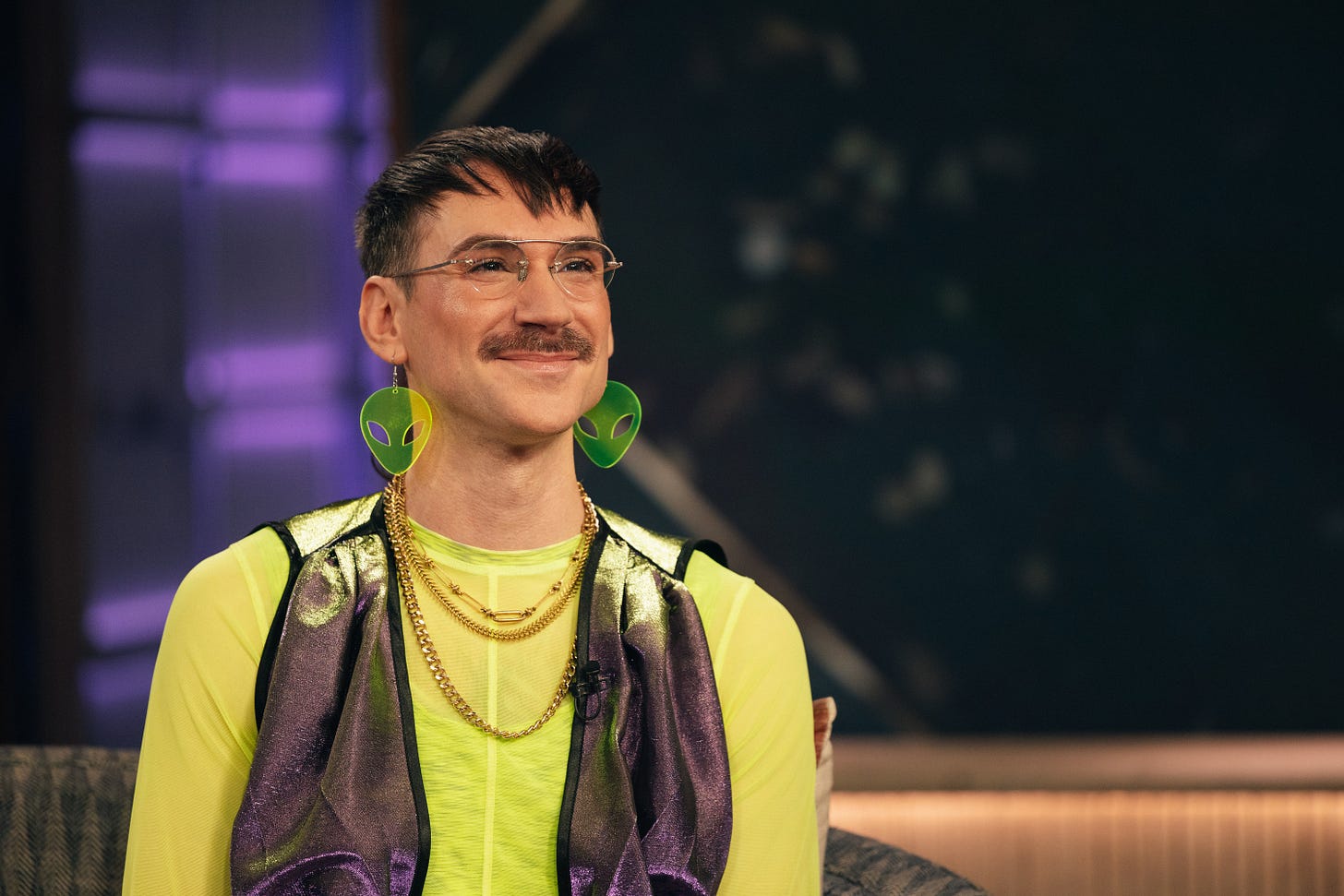 Kevin Gotkin, a white person with a mustache, is smiling from the couch on the set of The Kelly Clarkson Show. They are wearing glasses, big neon green earrings in the shape of an alien’s head, and gold necklaces over a neon green and shimmering purple outfit that looks like something out of the Ozdust Ballroom from Wicked.