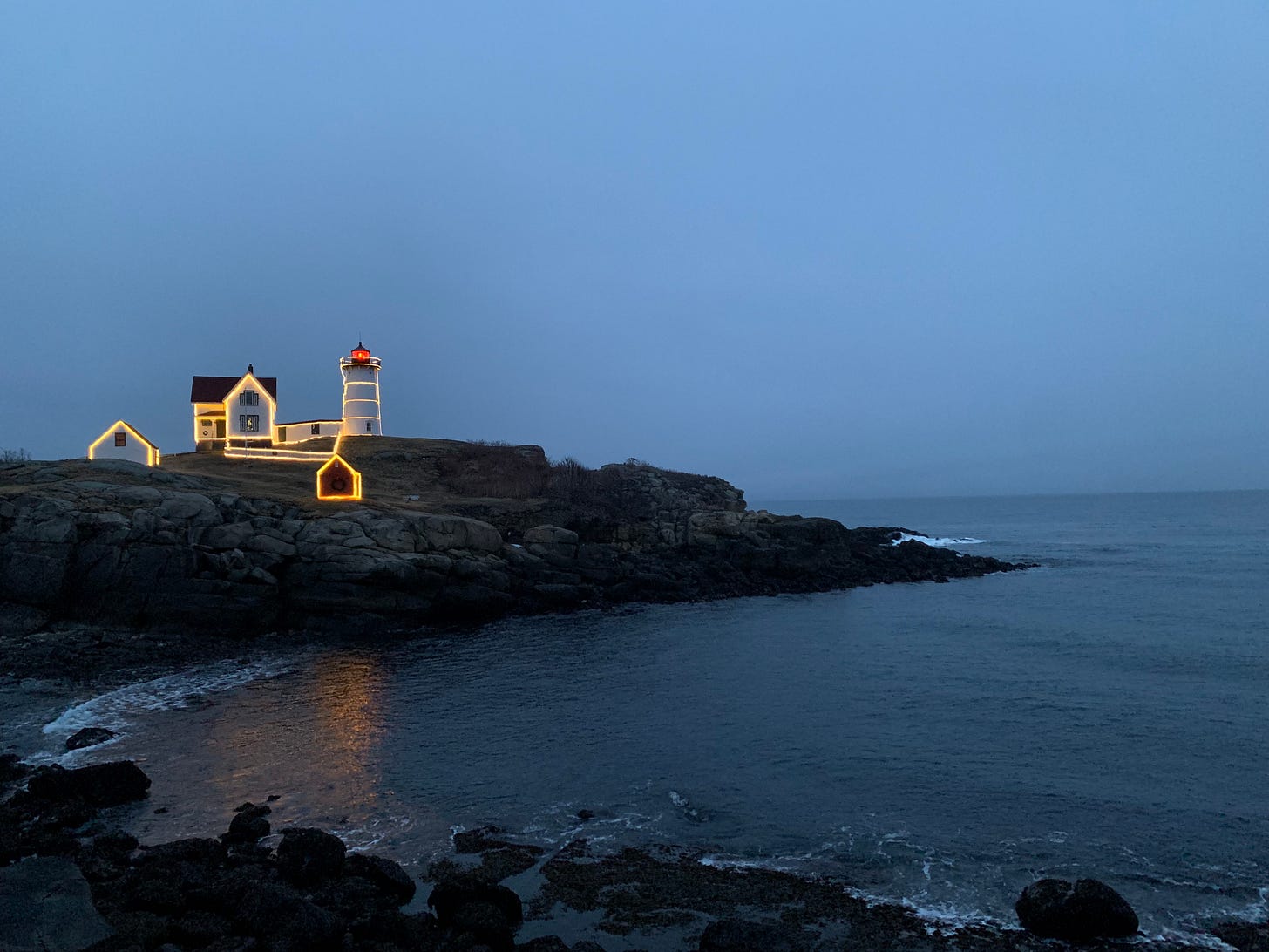 a lighthouse is decked with christmas lights and it is on a rocky bluff at dusk in maine.