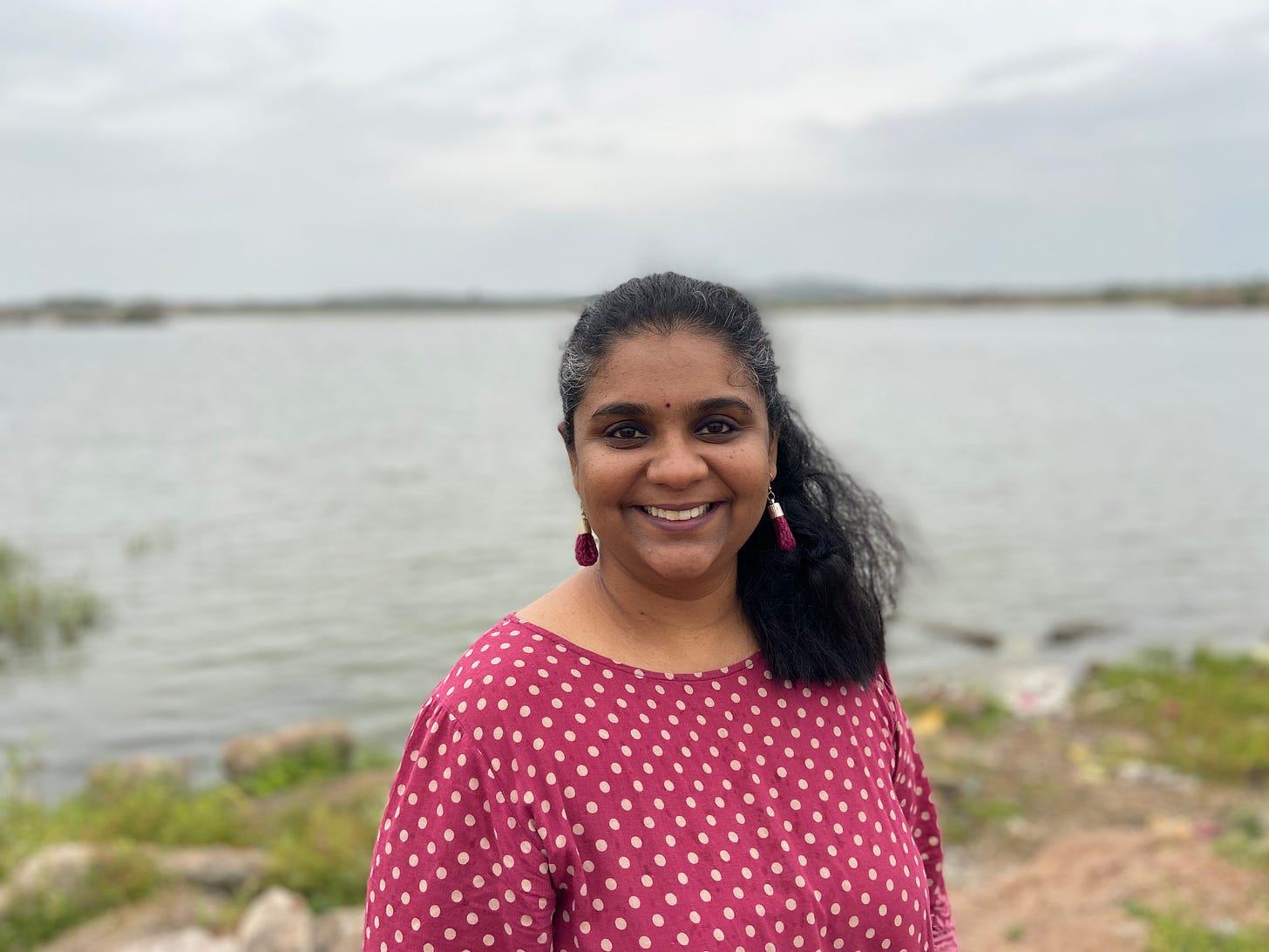 Standing on a lake shore, DVL Padma Priya, an Indian woman, has wavy black hair with grey streaks, tied in a ponytail. She is wearing a magenta dress with big white polka dots, matching bindi, and earrings, and smiling into the camera. 