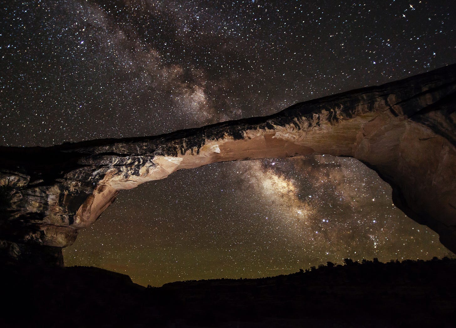 A bright band of stars crossed a dark sky full of stars with a geologic bridge formation in the foreground.