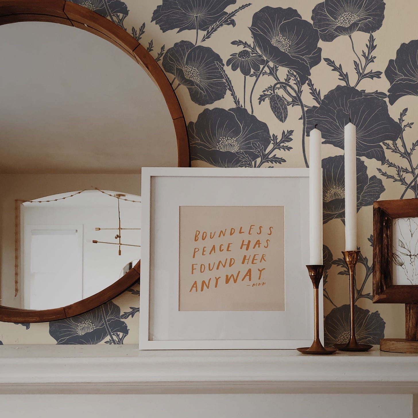 A mockup of the pattern on wallpaper. The image features a round wooden mirror above a mantel, upon which a framed art print and two candle sticks sit, in front of a wall featuring the new floral wallpaper pattern.