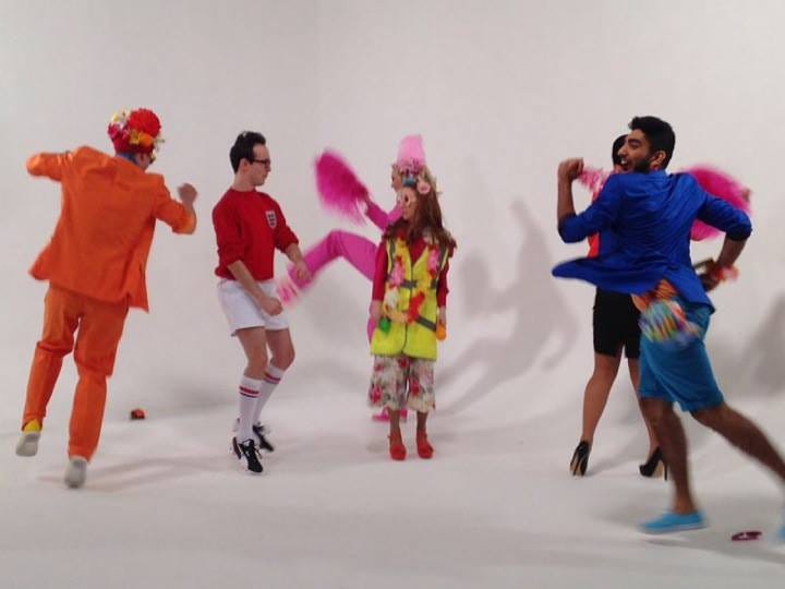 6 performers in brightly coloured clothing dance around against a white background.  