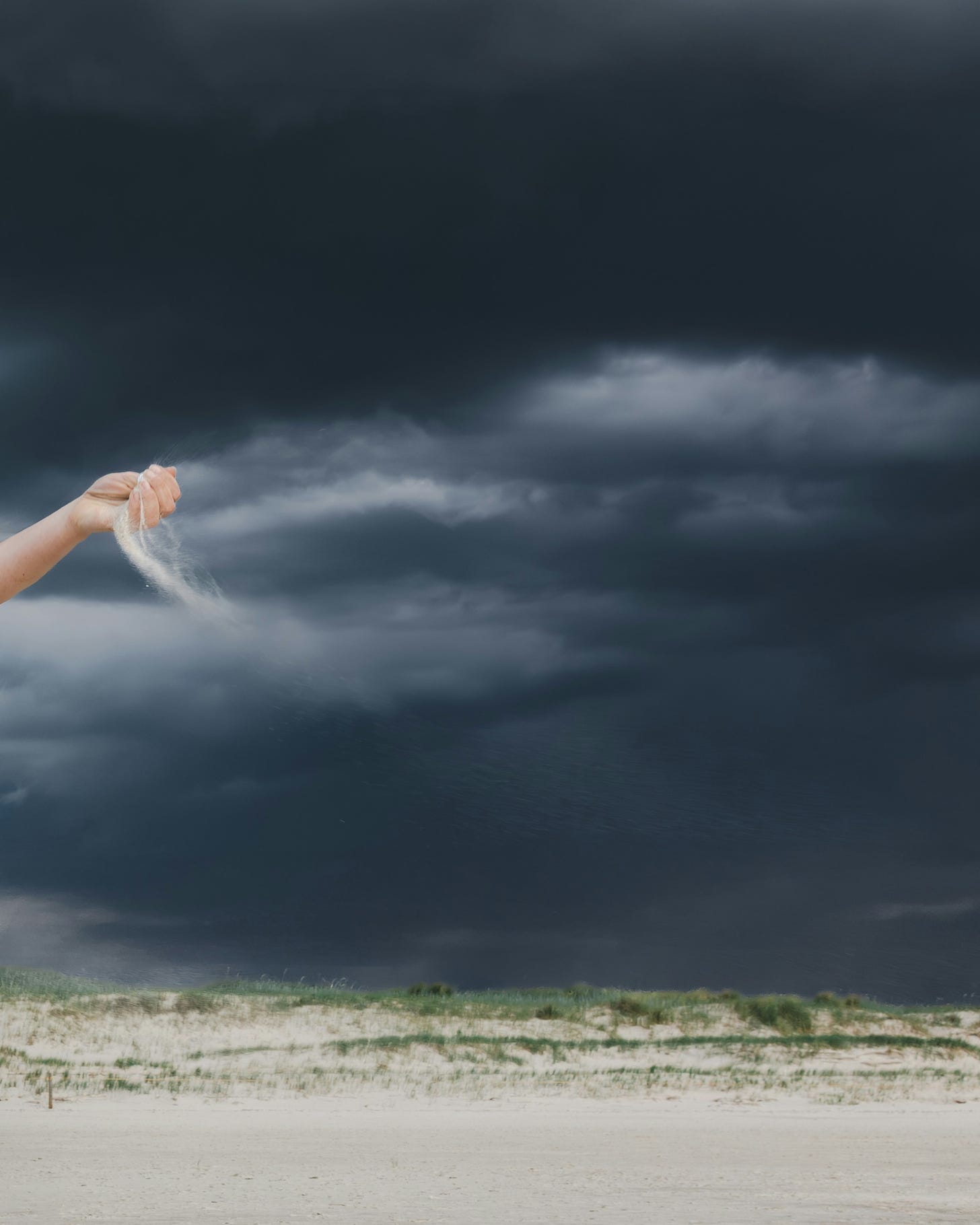 A white hand holds a wisp of white cloud against a stormy grey sky, letting the wisp disintegrate into the wind. 