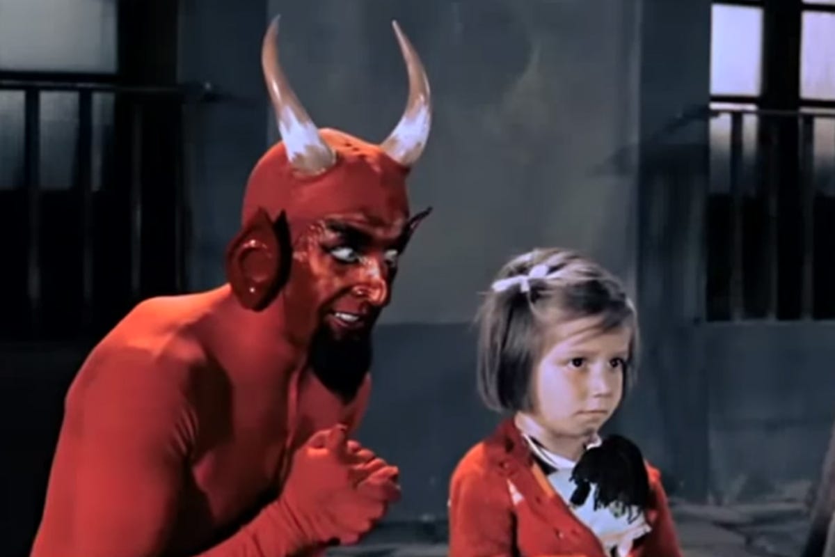 a long horned, long eared, beared, red devil leans over a child's shoulder as she clutches a doll.