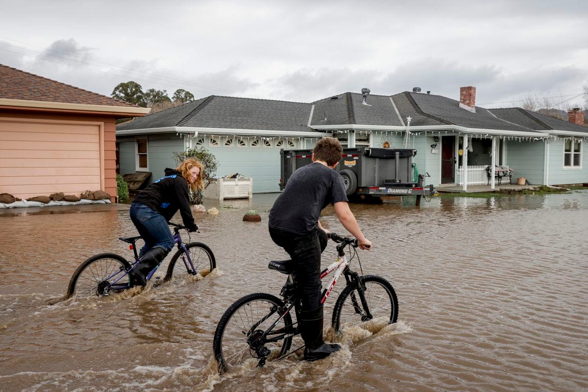 A boy and girl ride their bides through foot-deep water flooding a street, encroaching on porches and garages. 