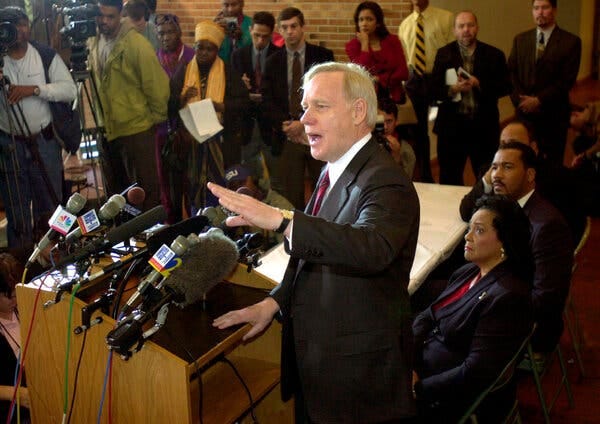 William Pepper, a white-haired man in a dark suit and a red tie, stands in front of a bank of microphones and gestures as he speaks. Members of the Rev. Dr. Martin Luther King Jr.’s family sit behind him.