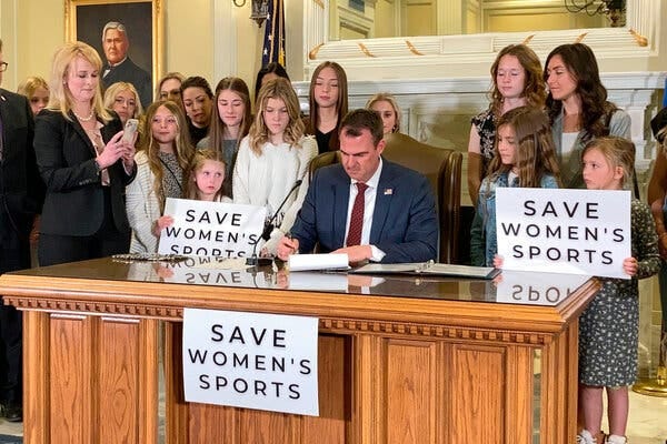 Gov. Kevin Stitt sits at a wooden desk signing a bill; behind him are several young female athletes, some with signs that read, “Save women’s sports.”