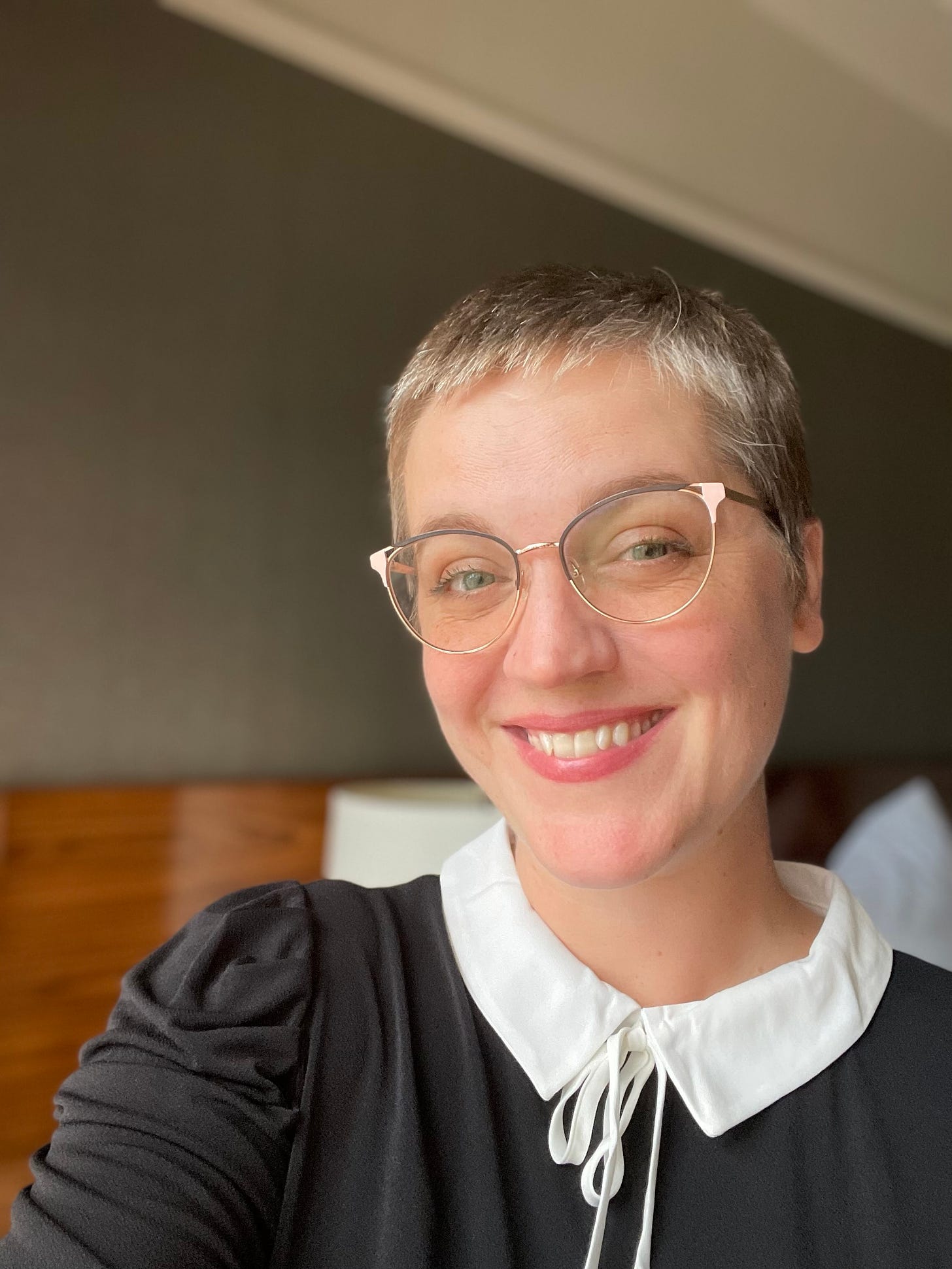 a white person socialied as female with super short hair is smiling at the camera.They are waring gold rimmed glasses and a tiny bit of makeup. They have on a black shirt with a white peter-pan collar, and sort of look like a quaker minister. 
