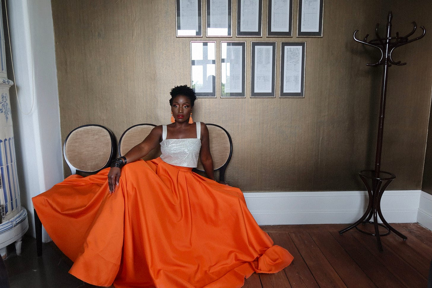 Lovette Jallow seated upright in an orange gown under framed writings, embodying resolve and reflection.