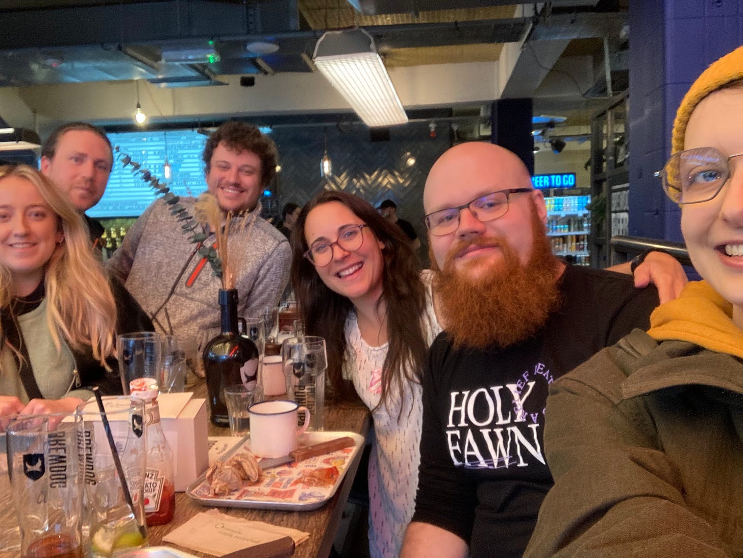 Selfie of 6 people in a bar celebrating Michaela passing her viva. Everyone looks pleased to be there except Marvin who is not quite posed for the photo so looks like he wants to be anywhere else.