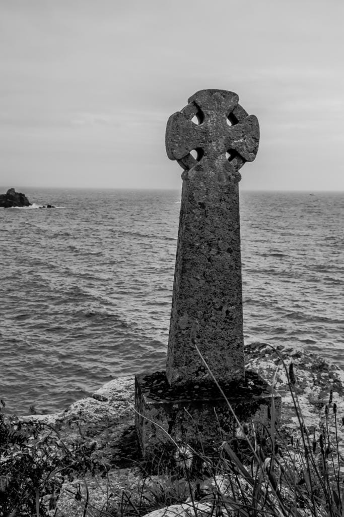 A black-and-white photo of a stone Celtic-style cross on a cliff with the sea beyond.