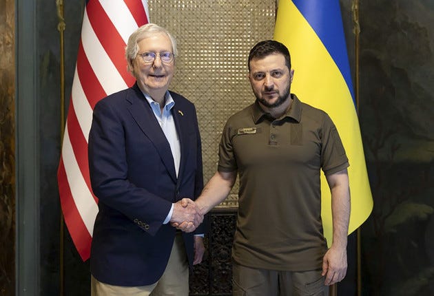 Volodymyr Zelenskyy and Mitch McConnell shake hands in front of American and Ukrainian flags. 