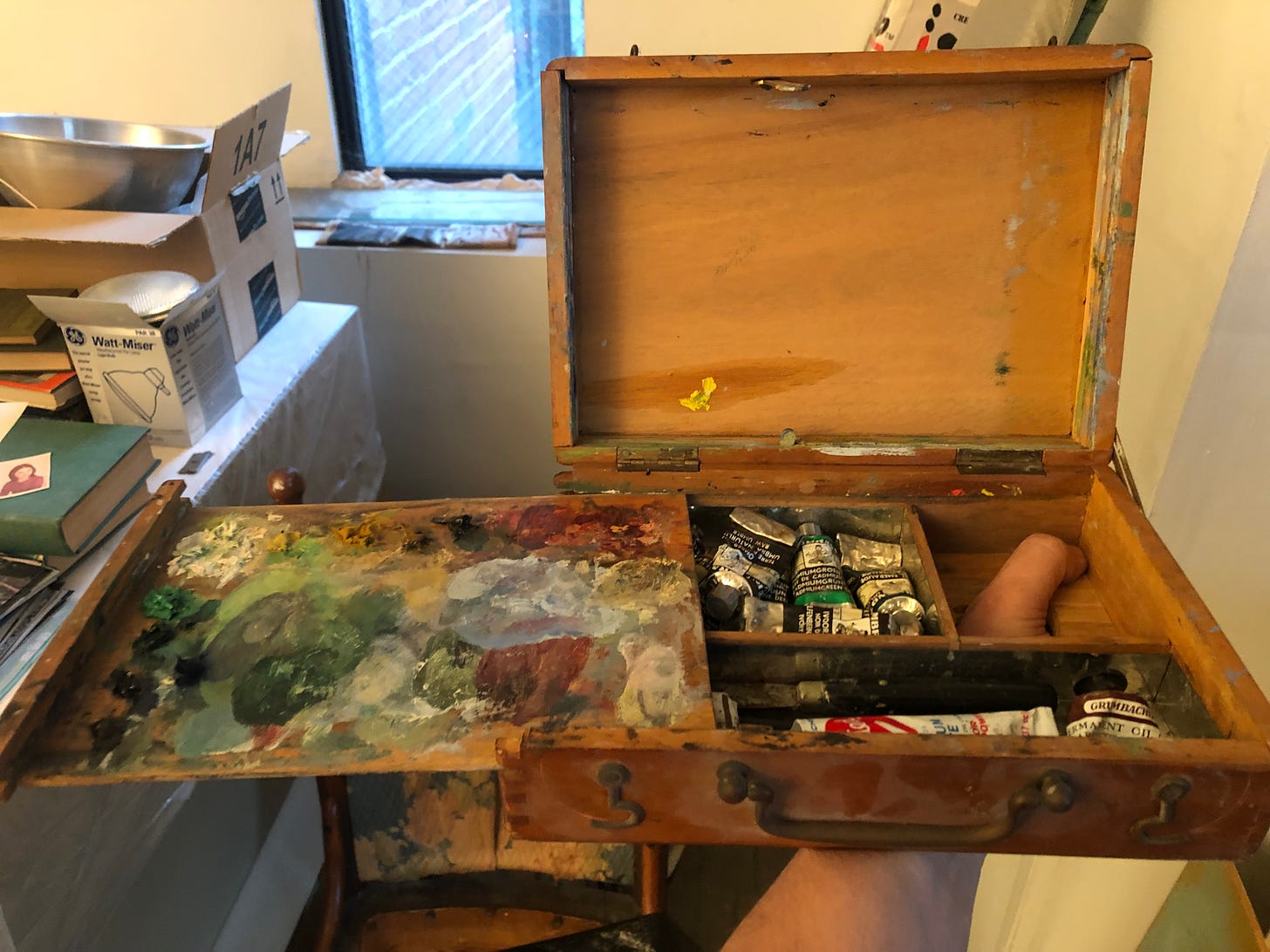 A wooden box with an attached palette slid sideways to reveal tubes of paint and a thumb gripping through a hole in the base of the box. There is a narrow window behind and studio clutter to the left.