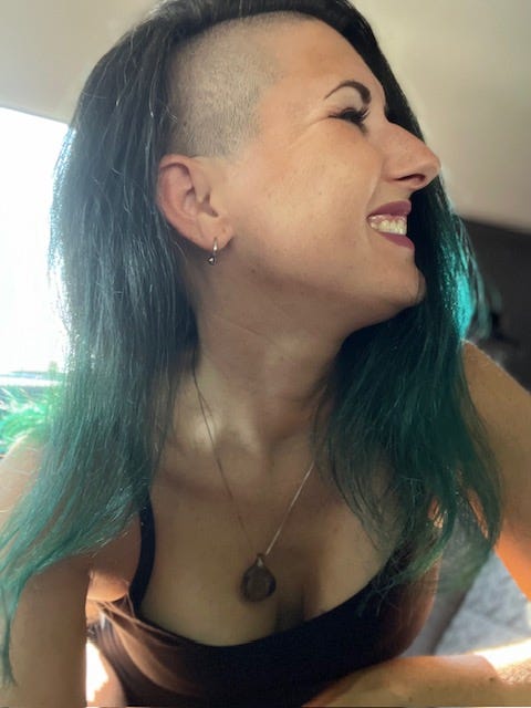 Recent photo of a happy and smiling Lyric, with long green hair and shaved sides, looking off to the right of the screen.