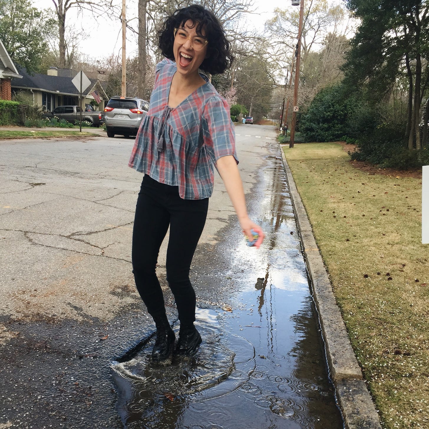 A picture of the author, a Japanese American with a curly bob, jumping in a puddle on a dreary day.