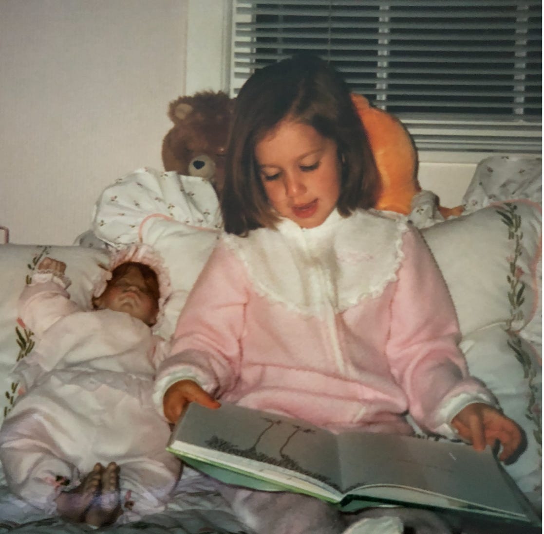 A child reading a book on a bed with a doll

Description automatically generated