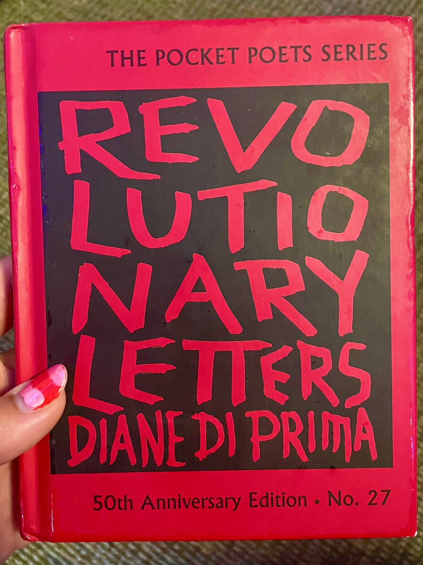 Me holding "Revolutionary Letters" by Diane di Prima. The cover is red with a black box. The title and author are in the black box in red font.