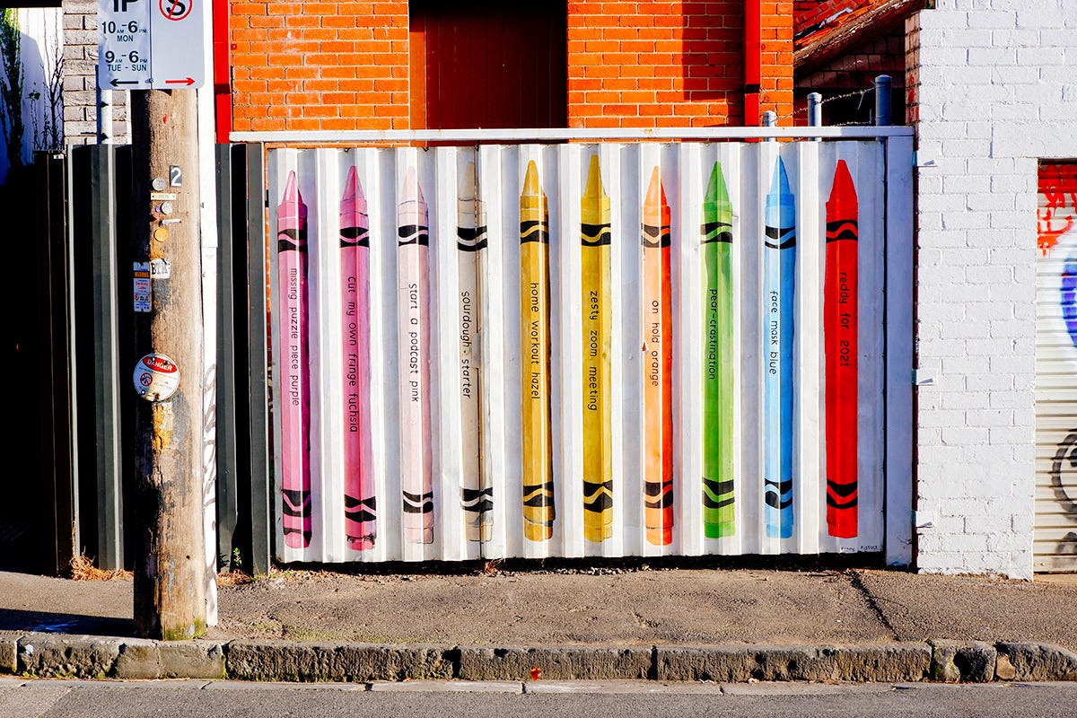 A wall in a street. On it art, painting of a box of crayons. There are funny things written on the crayons.