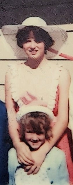 The image above shows a teenage Heidi standing in a pink Easter dress and bonnet with her hands around the shoulders of five-year-old Michele who is wearing a baby-blue Easter dress, sweater and bonnet. They are both smiling at the camera.